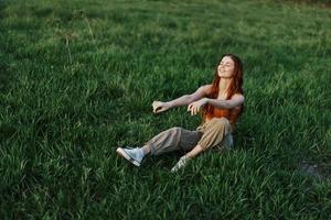 The redheaded woman sits in the park on the green grass wearing an orange top, green pants, and sneakers and looks out at the setting summer sun photo