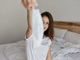 happy woman with medical mask in hand sitting on bed in a light room photo