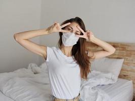 cheerful woman in a medical mask holds her hands near her face and sits on the bed indoors photo