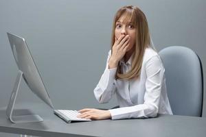 Shocked pretty blonde businesswoman worker freelancer using desktop computer close mouth by hand sitting at workplace in gray modern office. Remote Job And Career Profession Concept. Copy space photo