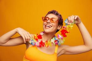 retrato de un mujer con un floral hawaiano guirnalda alrededor su cuello es teniendo divertido bailando y sonriente en brillante ropa en un naranja antecedentes vistiendo Gafas de sol, estilo de vida fiesta hawaiano estilo foto