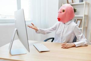 enojado mujer de negocios en rosado pescado máscara gritando a escritorio furioso después computadora software choque error a lugar de trabajo en moderno oficina. loco director trabajo en línea en financiero corporación. Copiar espacio foto
