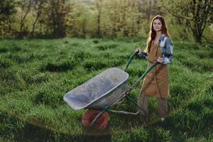 A happy woman with a cart works in her country home in the countryside against a backdrop of green grass and sunset sun photo