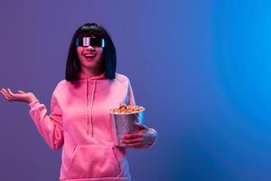 Laughing awesome brunet woman in pink hoodie trendy specular sunglasses with popcorn spread hand look aside posing isolated in blue violet color light background. Neon party Cinema concept. Copy space photo