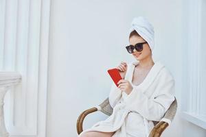 un mujer en un blanco túnica se sienta en un silla con un rojo teléfono en oscuro lentes aquí o en el Mañana foto