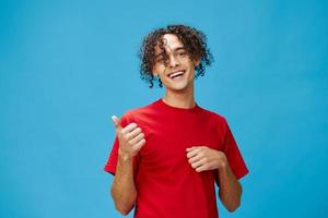 Happy cheerful positive curly tanned Caucasian guy in basic red t-shirt show thumbs up posing isolated on over blue background. Lifestyle and Emotions concept. Good offer with copy space for ad photo