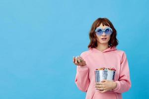 Evening movie concept. Confused cute redhead lady in pink hoodie sunglasses with popcorn posing isolated on blue studio background. Copy space Banner. Fashion Cinema. Entertainment offer photo