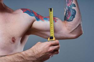 guy measures arm muscles with centimeters on a gray background and a multicolored tattoo bodybuilder fitness photo