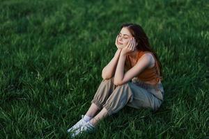 A beautiful girl relaxing in nature sitting in pants and top on the green grass in the rays of the setting summer sun. The concept of health and care for the body and the environment photo