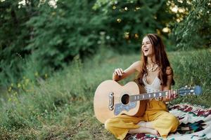 joven hembra hippie artista obras de teatro guitarra y canta canciones en Respetuoso del medio ambiente ropa sentado en el suelo fuera de en naturaleza en el otoño mirando fuera a el puesta de sol foto