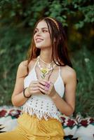 Young hippie woman sitting in nature on a plaid with a flower in her hands and looking at the camera smiling sincerely in eco-clothing in autumn in the sunset photo