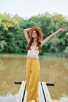 un joven mujer en un hippie Mira y eco-vestido viajes en naturaleza por el lago vistiendo un sombrero y amarillo pantalones en el otoño puesta de sol foto