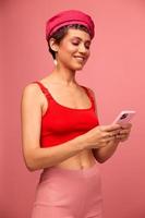 Young woman blogger with colored pink hair and short haircut flipping through the screen of the phone and typing a message with a smile in stylish clothes on a pink background photo