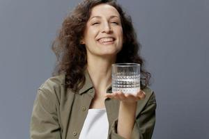 Cheerful adorable curly beautiful woman in casual khaki green shirt holds suggests glass of water posing isolated on over gray blue background. Healthy lifestyle. Water is Life concept. Copy space photo