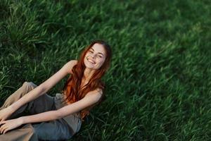 contento mujer sonriente hermosamente y mirando arriba a el cámara sentado en Fresco verde césped en el verano Brillo Solar foto