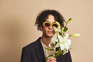 Funny cute handsome tanned curly man in trendy sunglasses hold lilies near face posing isolated on over beige pastel background. Fashion New Collection offer. Retro style concept. Free place for ad photo
