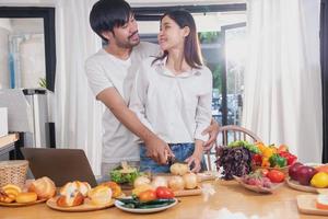 Young Asian couple cooking with fruits and vegetables and using laptop in the kitchen To cook food together within the family happily, family concept. photo