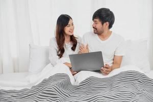 Young happy asian couple using laptop in bed watching movie, romantic time to enhance family relationship, family concept. photo