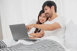 Young happy asian couple using laptop in bed watching movie, romantic time to enhance family relationship, family concept. photo