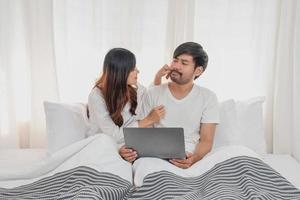 Young happy asian couple using laptop in bed watching movie, romantic time to enhance family relationship, family concept. photo