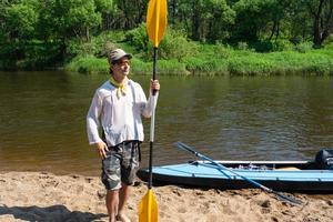 un hombre con un kayac paleta para canotaje soportes en el río banco. deporte agua caminata, un verano aventura. Respetuoso del medio ambiente y extremo turismo, activo y sano estilo de vida foto