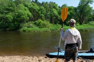 un hombre con un kayac paleta para canotaje soportes en el río banco. deporte agua caminata, un verano aventura. Respetuoso del medio ambiente y extremo turismo, activo y sano estilo de vida foto