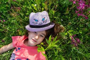 Portrait of a child in a hat with his face covered in summer lying in the grass and wildflowers. Hat with eyes and ears like a cat, summer time, freedom photo