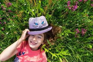 Portrait of a child in a hat with his face covered in summer lying in the grass and wildflowers. Hat with eyes and ears like a cat, summer time, freedom photo