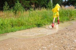A girl in red rubber boots and a yellow raincoat runs through puddles after a rain in the village. Summer time, freedom, childhood photo