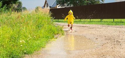 A girl in red rubber boots and a yellow raincoat runs through puddles after a rain in the village. Summer time, freedom, childhood photo