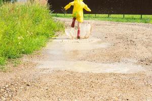 un niña en rojo caucho botas y un amarillo impermeable carreras mediante charcos después un lluvia en el aldea. verano tiempo, libertad, infancia foto