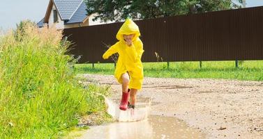 A girl in red rubber boots and a yellow raincoat runs through puddles after a rain in the village. Summer time, freedom, childhood photo