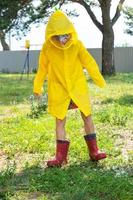 un niña en rojo caucho botas y un amarillo impermeable carreras mediante charcos después un lluvia en el aldea. verano tiempo, libertad, infancia foto