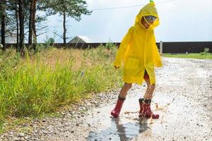 A girl in red rubber boots and a yellow raincoat runs through puddles after a rain in the village. Summer time, freedom, childhood photo