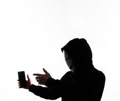 hacker anónimo y mascarilla con smartphone en la mano. hombre con camisa de capucha negra sosteniendo y usando un teléfono móvil con fondo blanco. representan el concepto de piratería informática o robo de datos personales foto