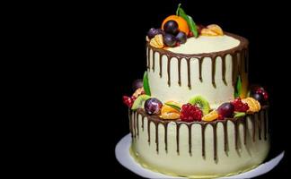 Beautiful two-tier cake poured with chocolate and decorated with fresh fruits on a black background. photo