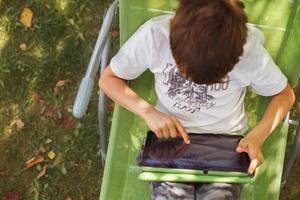 boy sitting with a tablet in the garden. the boy lies on a deck chair and plays on the tablet. child with a gadget photo