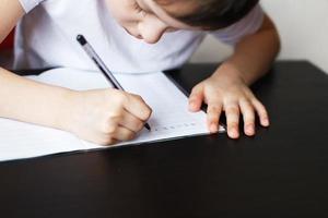 el chico se sienta a el mesa y escribe en un cuaderno. niño se sienta y lo hace deberes en un blanco antecedentes foto