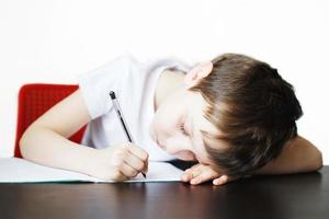 el chico se sienta a el mesa y escribe en un cuaderno. niño se sienta y lo hace deberes en un blanco antecedentes foto