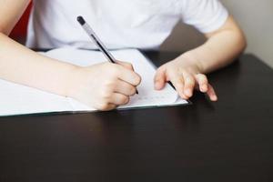 el chico se sienta a el mesa y escribe en un cuaderno. niño se sienta y lo hace deberes en un blanco antecedentes foto