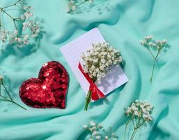 A bouquet of gypsophilia, a red heart made of sequins and a folded white sheet of paper on a textile background, a note. View from above photo