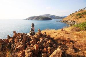 Beautiful Pyramid shape of stones arranged with Buddha statue in zen on rock mountain in seascape of sunset and sea horizon with Dry grass field on Phrom Thep Cape in Phuket island, Thailand. photo