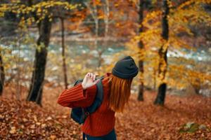 woman hiker with backpack in autumn forest near mountain river and fallen leaves photo