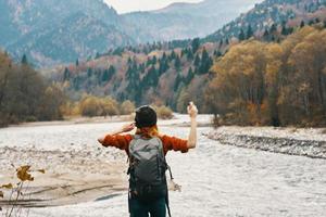 a traveler in a sweater hat with a backpack gestures with her hands and relaxes on the river bank in the mountains photo