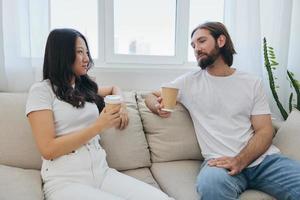 un hombre y un mujer sentado a hogar en el sofá en blanco elegante camisetas y chateando alegremente sonriente y riendo a hogar. masculino y hembra amistad foto