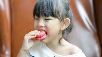madre entrega rojo con sabor a rosa macaron galletas a asiático niña de cerca. linda y alegre a comer deliciosamente en un cafetería. tarde ligero. adorable pequeño niña sentado en café y disfrutando delicioso video