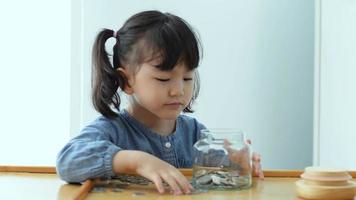 ahorro dinero. contento asiático niño niña salvar dinero por poniendo monedas en vaso frasco, concepto salvar dinero video