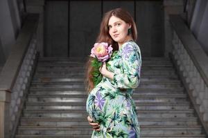 hermosa embarazada mujer con un flor en un antecedentes de gris escaleras. foto