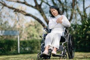 asiático antiguo mujer sentado en un silla de ruedas al aire libre en el parque tener dolor en el brazos, muñecas y cuerpo en Dom ligero foto