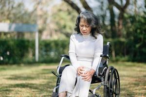 asiático antiguo mujer sentado en un silla de ruedas al aire libre en el parque tener dolor en el brazos, muñecas y cuerpo en Dom ligero foto